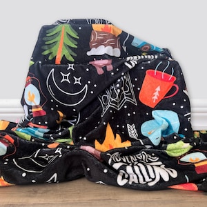 May include: A black fleece blanket with a camping theme. The blanket features illustrations of a campfire, a lantern, a mug, a moon, stars, and the text "Adventure is Calling".