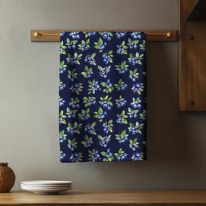 May include: A navy blue kitchen towel with a repeating pattern of watercolor blueberries and green leaves. The towel is hanging on a gold-colored rod. The towel is made of a soft, absorbent material.