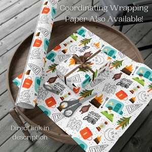 May include: White wrapping paper with a camping theme, featuring illustrations of campfires, bears, and travel trailers. A roll of paper and a wrapped gift box are visible, along with scissors and tape. The text "Explore" and "Adventure is Calling" are printed on the paper.