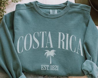 Peut inclure: Un sweat-shirt de couleur sarcelle avec l'inscription "COSTA RICA" en grand arc. En dessous, un dessin de palmier et le texte "EST. 1821". Le sweat-shirt a un col rond et des manches longues.