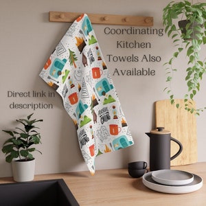 May include: White kitchen towel with a camping-themed pattern. The design includes illustrations of a camper, campfire, mountains, and mugs. Text on the towel reads "Adventure is Calling" and "Explore". Additional text says "Coordinating Kitchen Towels Also Available" and "Direct link in description".
