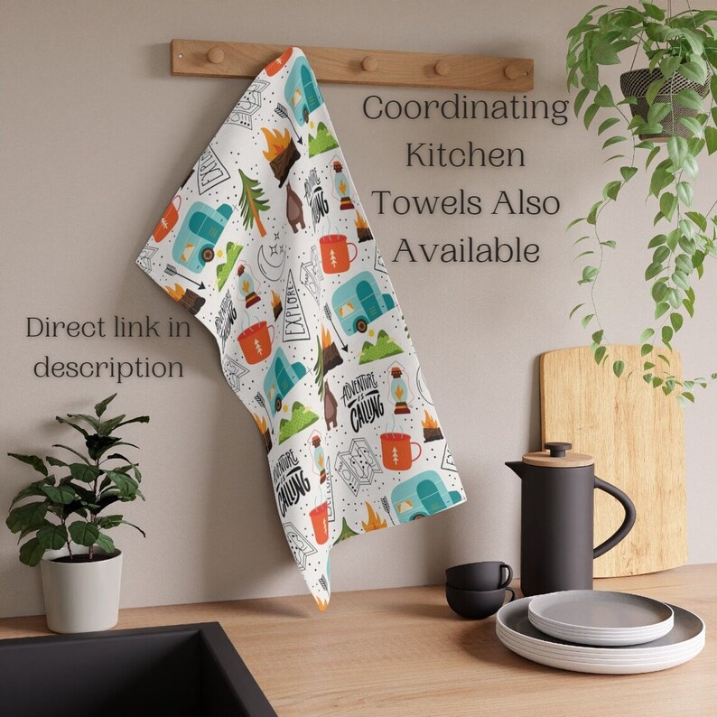 May include: White kitchen towel with a camping-themed pattern. The design includes illustrations of a camper, campfire, mountains, and mugs. Text on the towel reads "Adventure is Calling" and "Explore". Additional text says "Coordinating Kitchen Towels Also Available" and "Direct link in description".