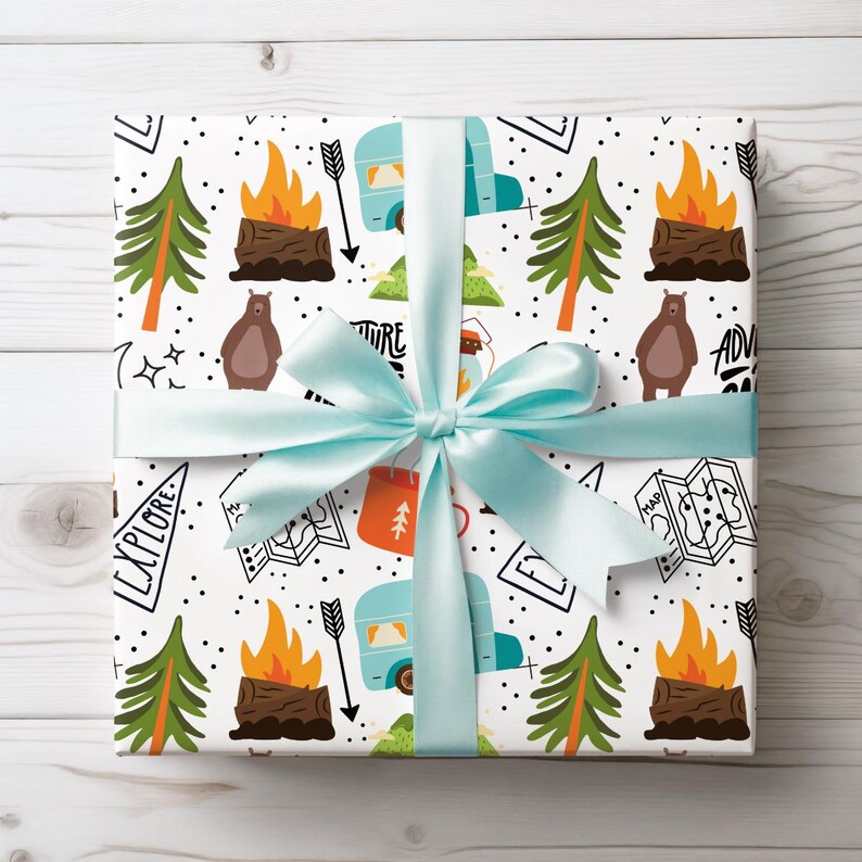 May include: A square gift wrapped in white paper with a camping-themed pattern, featuring illustrations of campfires, trees, bears, and a camper. A light blue ribbon is tied around the gift. The words "Explore" and "Adventure" are visible.
