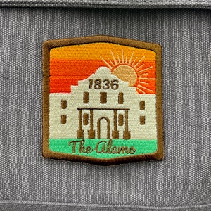 May include: A brown leather bag with a patch featuring a white silhouette of the Alamo with the text "1836 The Alamo" and a sunset graphic.