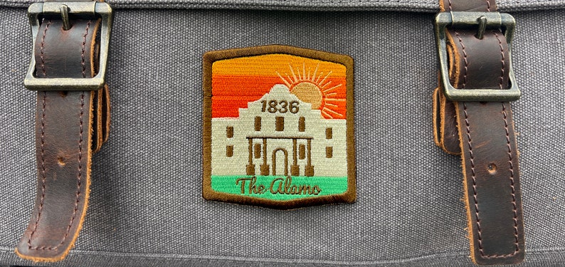 May include: A brown leather bag with a patch featuring a white silhouette of the Alamo with the text "1836 The Alamo" and a sunset graphic.