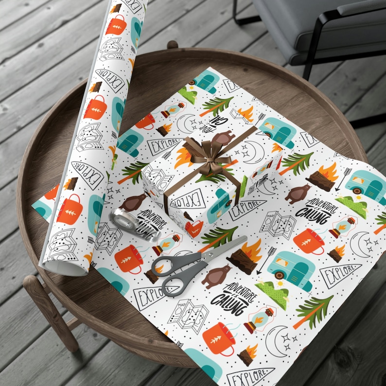May include: White wrapping paper with a camping theme, featuring illustrations of a camper, bear, campfire, and map. A wrapped gift with a brown ribbon sits on the paper. The words "Adventure Calling" and "Explore" are printed on the paper.