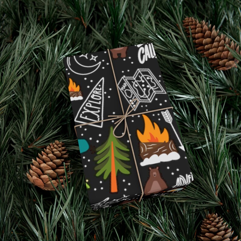 May include: Black wrapping paper with a camping theme, featuring illustrations of a campfire, trees, a bear, a map, and the word "EXPLORE". The paper is tied with twine and set against a backdrop of pine needles and pine cones.