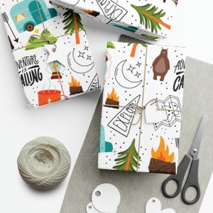 May include: Overhead shot of wrapped gifts with camping-themed paper. The white paper features illustrations of a camper, trees, a campfire, and the text "Adventure is Calling" and "Explore". The gifts are tied with twine and have white tags. A ball of twine, scissors, and tags are also visible.