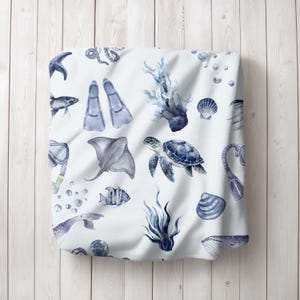 May include: White blanket with a blue watercolor pattern of marine life, including a sea turtle, fish, and coral. The design also features scuba gear, bubbles, and seashells. The blanket is displayed on a light wood background.