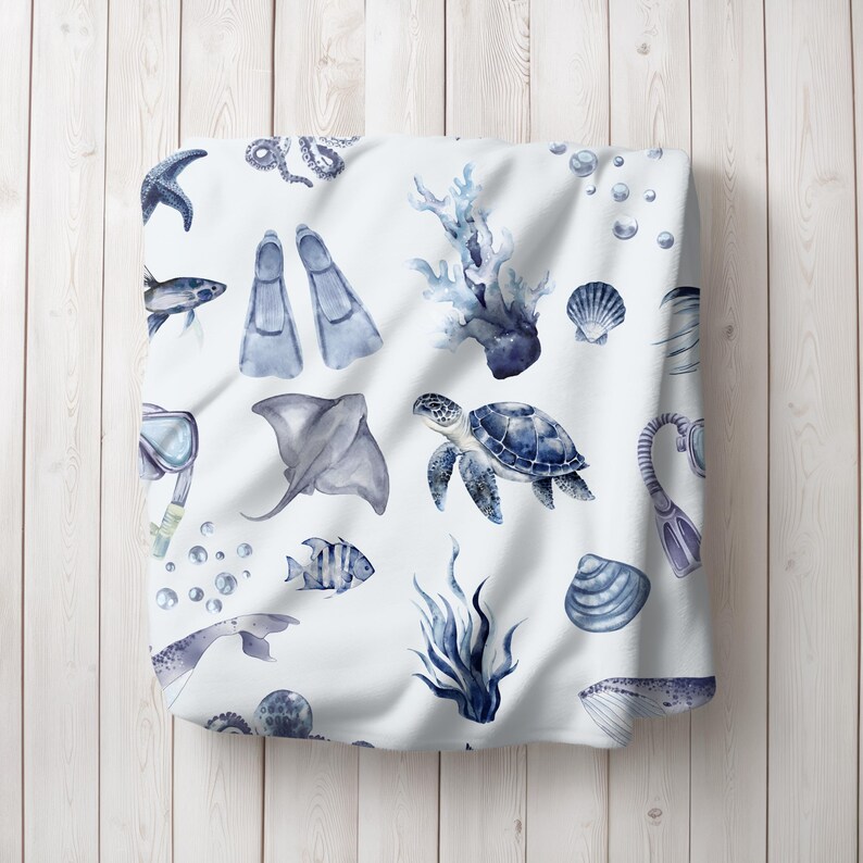 May include: White blanket with a blue watercolor pattern of marine life, including a sea turtle, fish, and coral. The design also features scuba gear, bubbles, and seashells. The blanket is displayed on a light wood background.