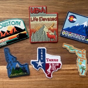 May include: A collection of embroidered patches featuring state and national park designs. The patches are colorful and feature various landscapes, including mountains, forests, and rivers. The patches are perfect for adding a touch of personality to backpacks, jackets, or other items.