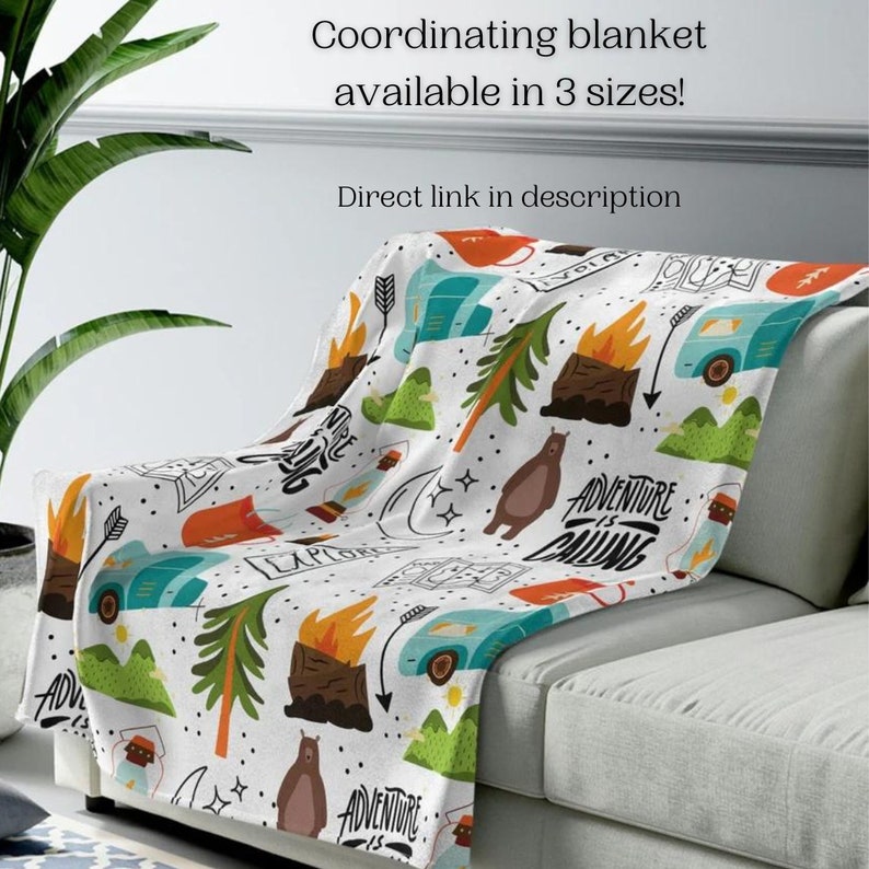 May include: A white blanket with a colorful camping-themed pattern, including bears, campfires, trees, and travel trailers. The text "ADVENTURE IS CALLING" is visible. The blanket is draped over a gray sofa. Text on the image says "Coordinating blanket available in 3 sizes!"