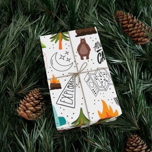 May include: White gift wrap with a camping-themed pattern featuring a bear, trees, a campfire, a map, and the words "Explore" and "Adventure". The gift is tied with twine and set against a backdrop of pine needles and pine cones.