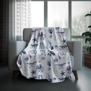 May include: A white throw blanket draped over a chair, featuring a repeating pattern of marine life in shades of blue. The design includes whales, octopuses, starfish, and scuba gear. The blanket is made of a soft material.