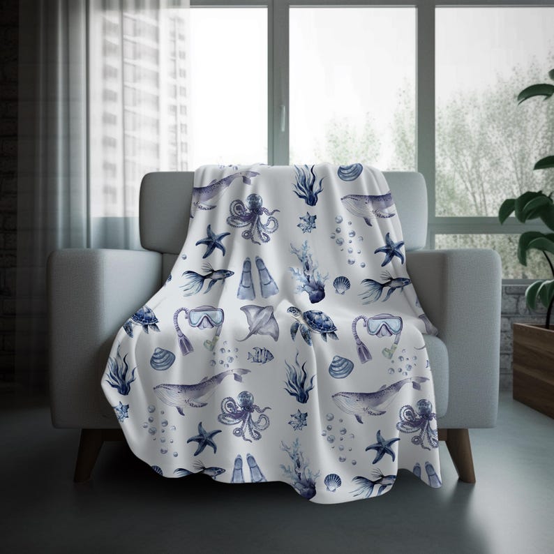 May include: A white throw blanket draped over a chair, featuring a repeating pattern of marine life in shades of blue. The design includes whales, octopuses, starfish, and scuba gear. The blanket is made of a soft material.