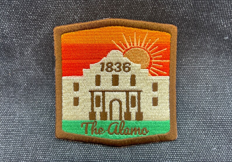 May include: A brown, orange, and green embroidered patch with a white building and a sun. The building has the text "1836" above the entrance and the text "The Alamo" below.