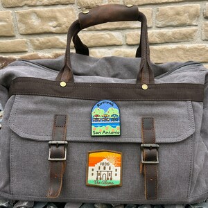 May include: A gray canvas travel bag with brown leather accents and two patches. The patches are rectangular and feature colorful illustrations of the Riverwalk and the Alamo in San Antonio, Texas.