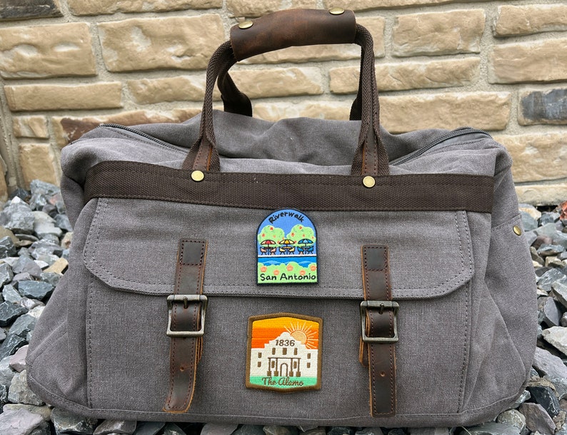 May include: A gray canvas travel bag with brown leather accents and two patches. The patches are rectangular and feature colorful illustrations of the Riverwalk and the Alamo in San Antonio, Texas.