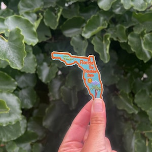 May include: A blue and orange embroidered patch shaped like the state of Florida. The patch says "Florida The Sunshine State" with a palm tree and sun graphic.