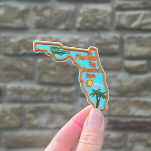 May include: A blue felt patch shaped like the state of Florida with an orange outline. The patch has an alligator, a sun, a cloud, and palm trees embroidered on it. The text "Florida The Sunshine State" is embroidered on the patch.