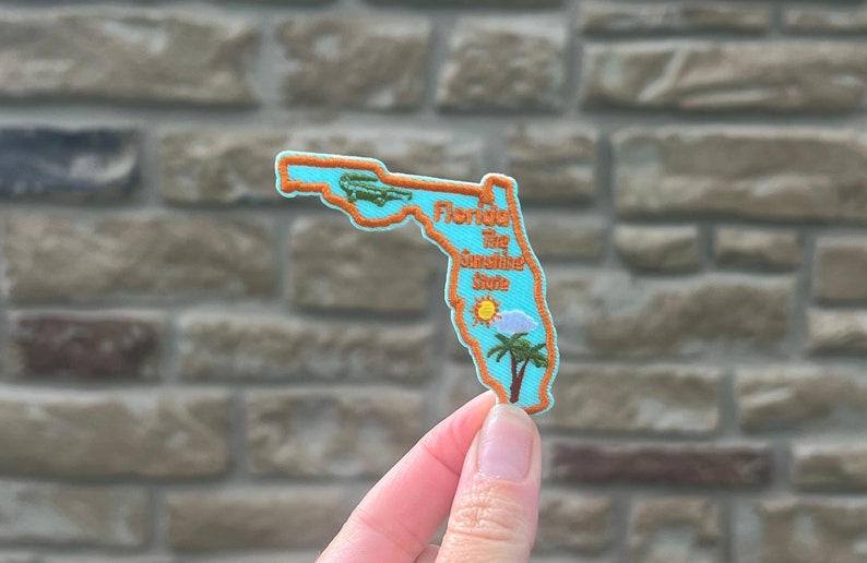 May include: A blue felt patch shaped like the state of Florida with an orange outline. The patch has an alligator, a sun, a cloud, and palm trees embroidered on it. The text "Florida The Sunshine State" is embroidered on the patch.