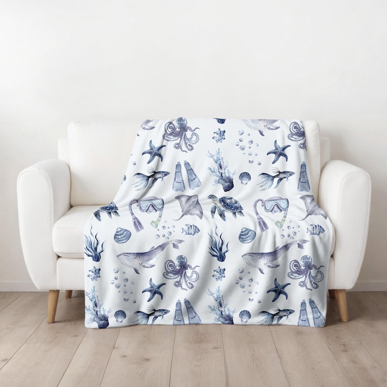 May include: A white throw blanket with a repeating pattern of blue watercolor sea creatures, including whales, octopuses, starfish, and fish. The blanket is draped over a white sofa. The design also includes scuba masks, flippers, and coral.