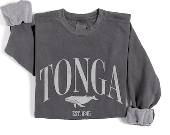 May include: A charcoal grey sweatshirt featuring the word "TONGA" in white, arched lettering. A white whale graphic and the text "EST. 1845" are below. The sleeves have grey cuffs. The sweatshirt is made of a soft material.