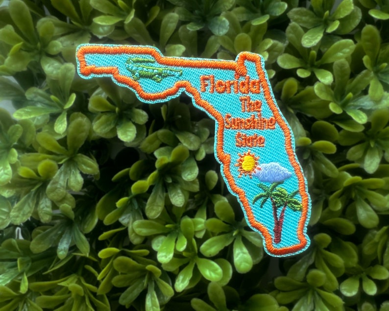 May include: An orange and blue embroidered patch shaped like the state of Florida. The patch features the text "Florida The Sunshine State" and a cartoon sun, cloud, and palm tree.