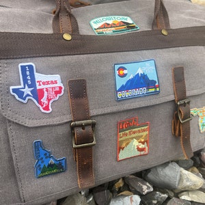 May include: A gray canvas travel bag with brown leather straps and buckles. The bag has several embroidered patches sewn on it, including patches for Texas, Colorado, Utah, and Yellowstone National Park.