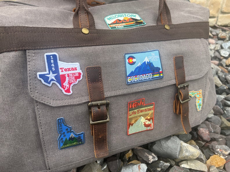 May include: A gray canvas travel bag with brown leather straps and buckles. The bag has several embroidered patches sewn on it, including patches for Texas, Colorado, Utah, and Yellowstone National Park.