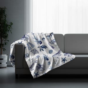 May include: A white throw blanket draped over a gray sofa, featuring a repeating pattern of marine life in shades of blue. The design includes octopuses, whales, sea turtles, starfish, and other sea creatures.