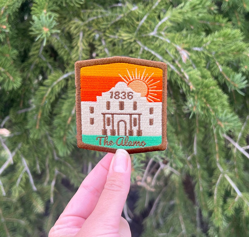 May include: A brown and green embroidered patch with a sunset and the Alamo building. The text "1836" and "The Alamo" are embroidered on the patch.