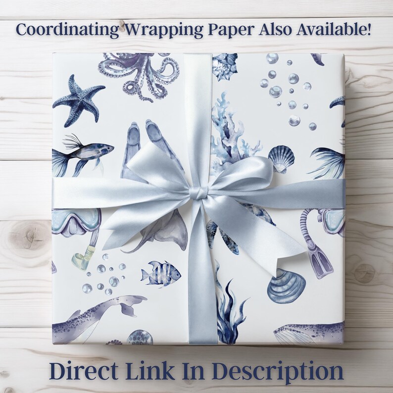 May include: White gift wrap with a blue ocean-themed design, featuring starfish, fish, and scuba gear. A light blue satin ribbon is tied around the package. Text on the wrapping paper reads: "Coordinating Wrapping Paper Also Available!" and "Direct Link In Description."