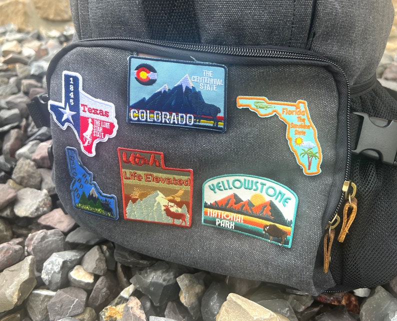 May include: A gray backpack with several colorful embroidered patches featuring state names and logos, including Texas, Colorado, Utah, Florida, and Yellowstone National Park.