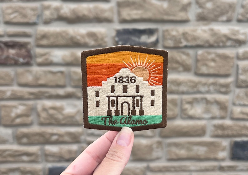 May include: A brown, beige, and green embroidered patch with a sunset scene and the Alamo building. The text "1836" and "The Alamo" are embroidered on the patch.