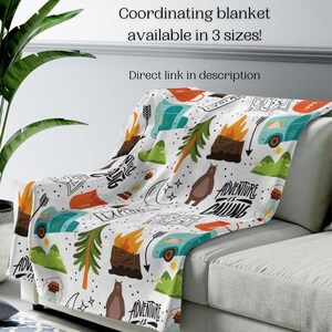 May include: A white blanket with a colorful camping-themed pattern, including bears, campfires, trees, and camper vans. The text "Adventure is Calling" is visible. The blanket is draped over a gray sofa. Text on the image says "Coordinating blanket available in 3 sizes!"