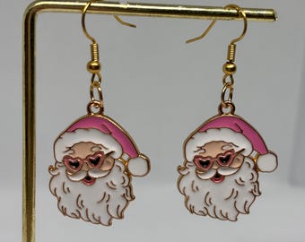 May include: A pair of gold-coloured Santa Claus earrings. Each earring features a Santa face with a white beard, pink hat, and heart-shaped sunglasses. The earrings are suspended from gold-coloured fish hook ear wires.