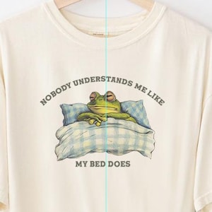 May include: Cream-colored t-shirt featuring a frog in bed with the text "NOBODY UNDERSTANDS ME LIKE MY BED DOES." The frog is green with a blue and white checkered pillow and blanket. The shirt is an original design by Fairly Stupid Shirts.