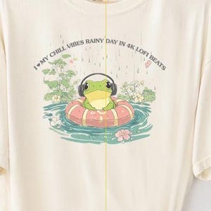 Op de afbeelding: Een crèmekleurig T-shirt met een groene kikker die een koptelefoon draagt en op een roze zwemband in een blauwe vijver drijft. De tekst op het shirt luidt "I ♥ my chill vibes rainy day in 4k lofi beats".