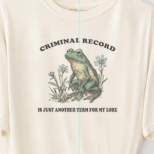 May include: A cream-colored t-shirt with a frog illustration and the text "Criminal Record is just another term for my lore".