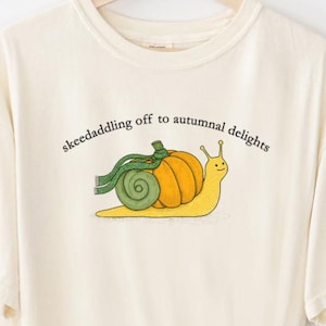 May include: Cream-coloured t-shirt with a cartoon snail wearing a pumpkin shell and green scarf. The text says "skeedaddling off to autumnal delights." An original design by Fairly Stupid Shirts.