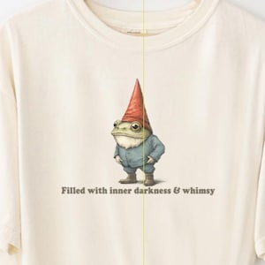 May include: Cream-colored t-shirt featuring a whimsical illustration of a frog dressed as a garden gnome, wearing a red hat and blue jacket. The text "Filled with inner darkness & whimsy" is printed below the frog. The shirt is an original design by Fairly Stupid Shirts.
