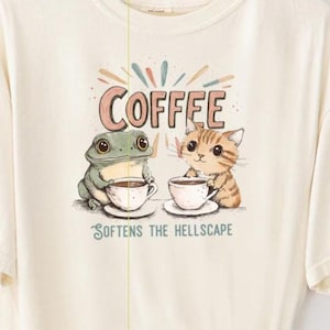 May include: A cream colored t-shirt with a graphic of a frog and a cat drinking coffee. The text "Coffee" and "Softens the Hellscape" are printed on the shirt.