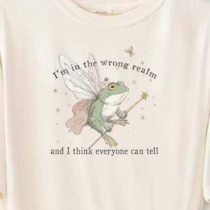 May include: Cream-colored t-shirt featuring a whimsical illustration of a frog with fairy wings, a crown, and a wand. The text "I'm in the wrong realm and I think everyone can tell" is printed above the frog. The shirt is an original design by FAIRLYSTUPIDSHIRTS.