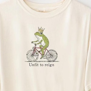 Può includere: T-shirt color crema con una rana verde con corona che va in bicicletta rossa. Il testo "Unfit to reign" è sotto la grafica. La maglietta è un design originale di Fairly Stupid Shirts.