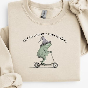 May include: A cream-coloured sweatshirt featuring the text "Off to commit tom foolery" and a cartoon frog in a wizard hat riding a scooter. The design is by Fairly Stupid Shirts.