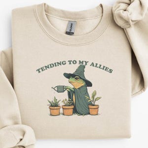 Puede incluir: Sudadera beige con una ilustración de una rana con sombrero y capa de mago regando plantas. El texto "TENDING TO MY ALLIES" está arriba. Diseño original de Fairly Stupid Shirts.