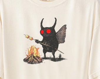 May include: A cream-colored t-shirt with a black and red illustration of a creature with horns and wings roasting a marshmallow over a campfire. The text "FairlyStupidShirts" is printed on the bottom right corner of the shirt.
