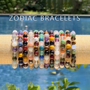 May include: A collection of colorful beaded bracelets, each featuring a different gemstone. The bracelets are stacked on a beige surface and are labeled "Zodiac Bracelets".