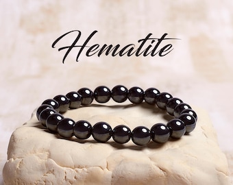 May include: A black hematite beaded bracelet on a light beige surface. The word "Hematite" is written in black cursive above the bracelet.
